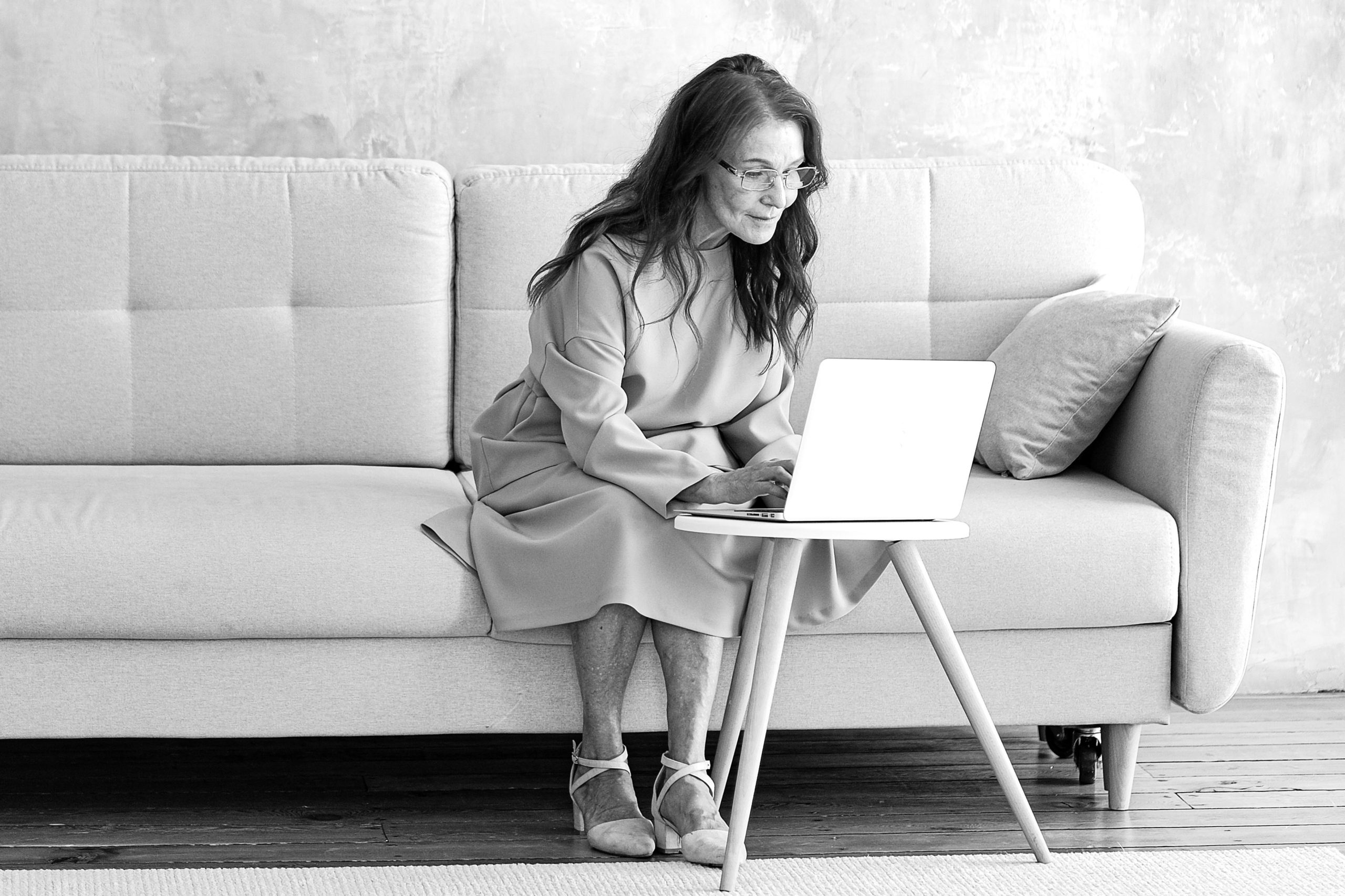 Woman in a dress sits on a sofa and works on a laptop that is on a small table.